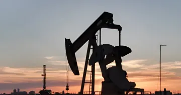 Silhouette of an oil pump jack against a sunset and drilling rig
