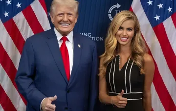 Дональд Трамп и молодая блондинка в темном платье стоят на фоне американских флагов и показывают большой палец вверх