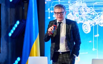 Глава Немецко-Украинской торговой палаты Райнер Перау выступает с микрофоном на фоне украинского флага