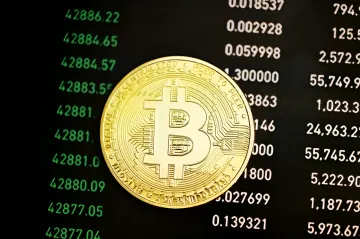 A gold bitcoin coin lies against the background of a black screen with green cryptocurrency trading charts
