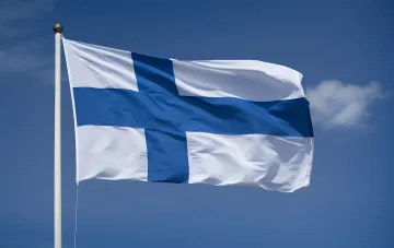 Die Flagge Finnlands weht an einem weißen Fahnenmast vor einem klaren blauen Himmel mit einer Wolke
