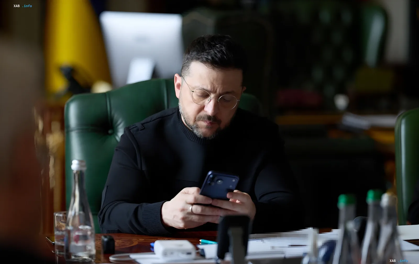 Президент Украины Владимир Зеленский в черном свитере в очках просматривает телефон в рабочем кабинете