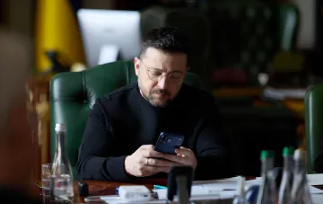Президент Украины Владимир Зеленский в черном свитере в очках просматривает телефон в рабочем кабинете