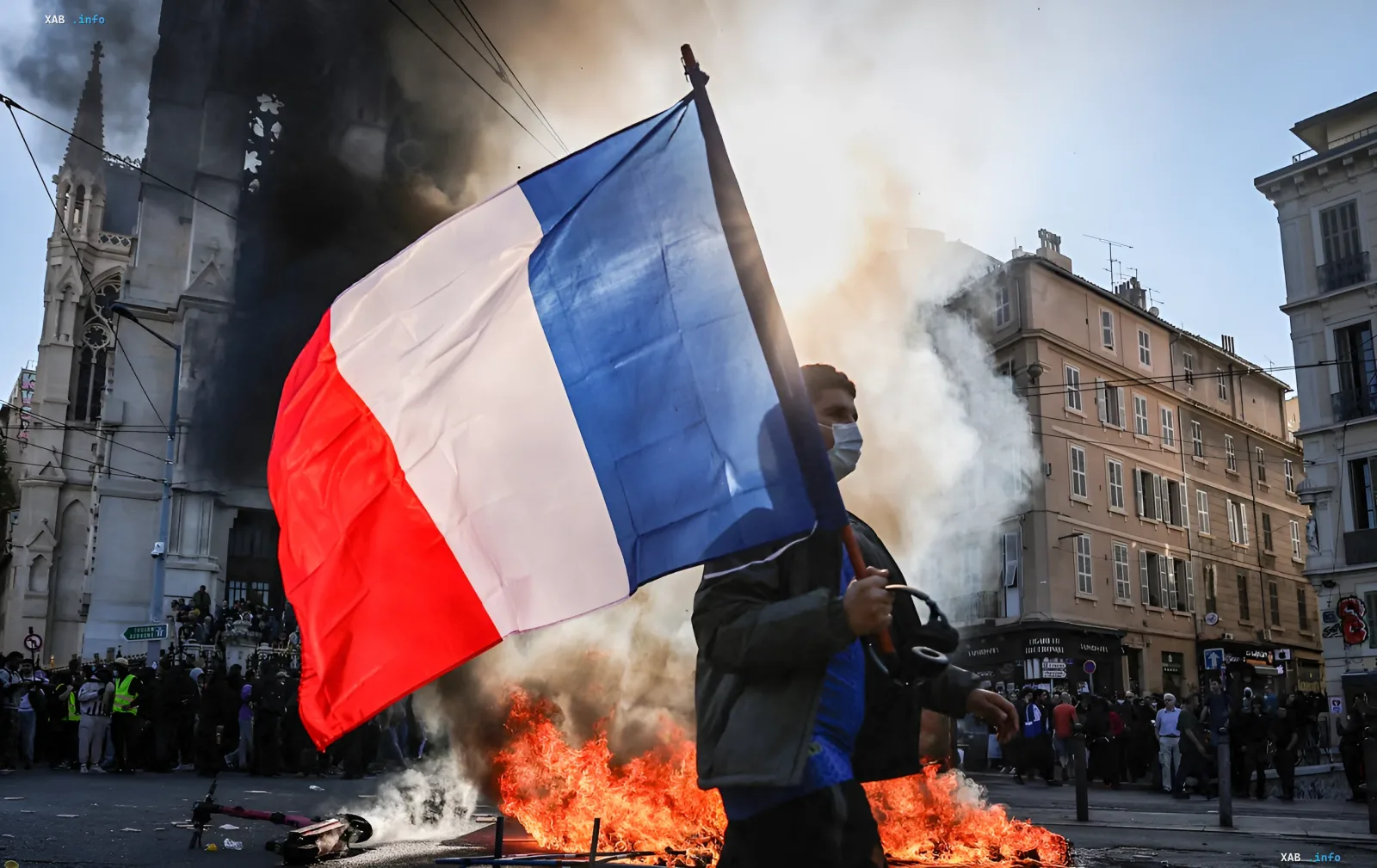 A man holding a French flag against the backdrop of a burning barricade and smoke during street riots