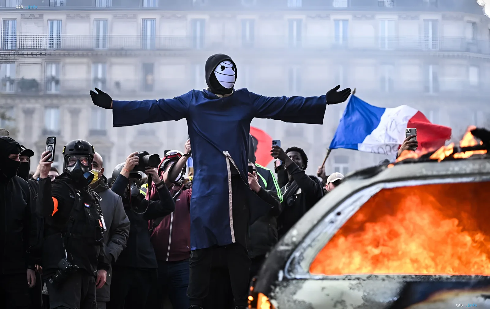 Protester in a mask and blue cloak with outstretched arms against the background of a burning car in Paris