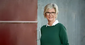 Frau mit Brille und grünem Pullover steht vor einer Wand