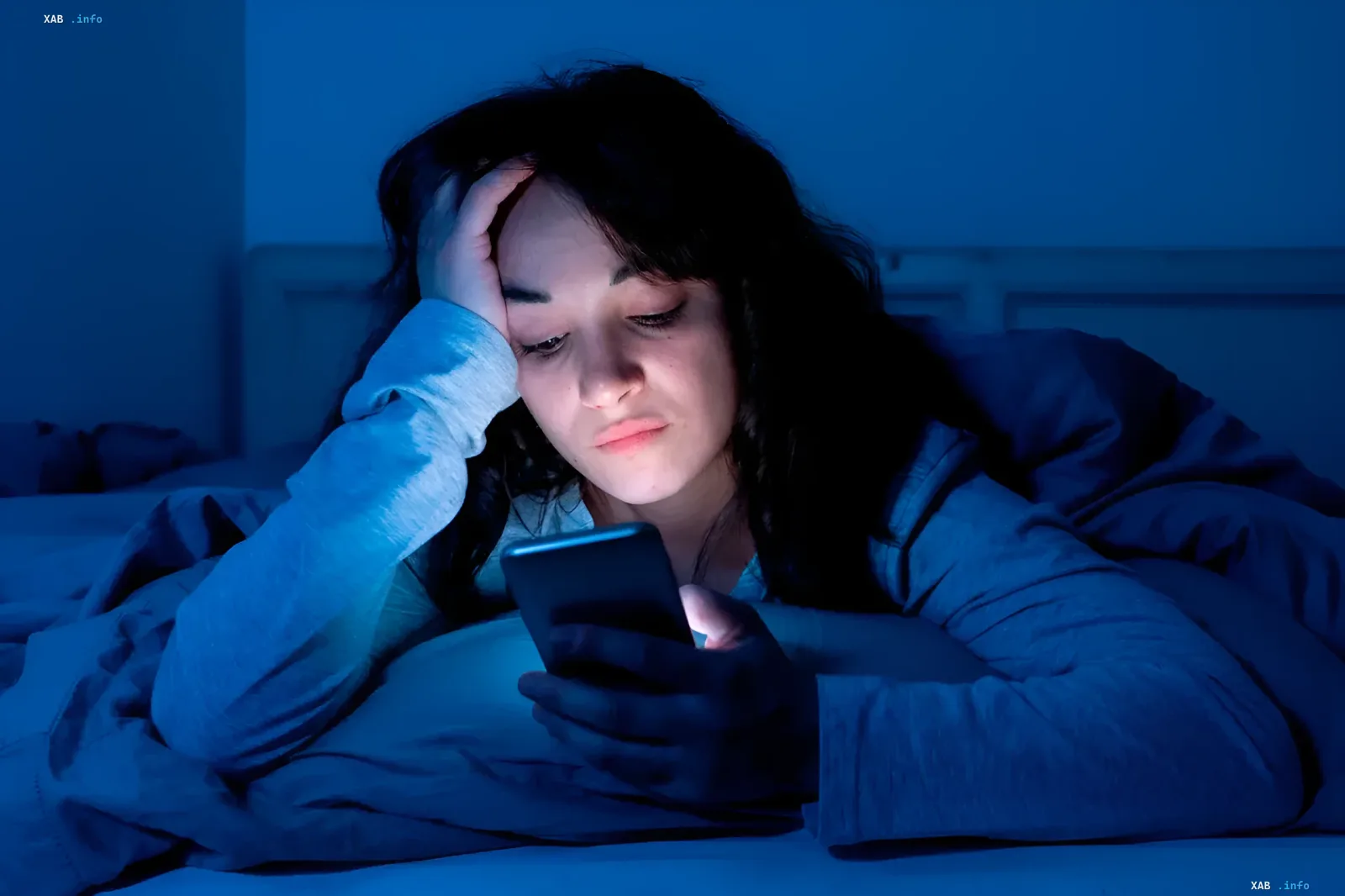 Eine Frau liegt im Dunkeln im Bett und blickt mit besorgtem Gesichtsausdruck auf ein Smartphone, dessen Bildschirm ihr Gesicht hell anstrahlt.