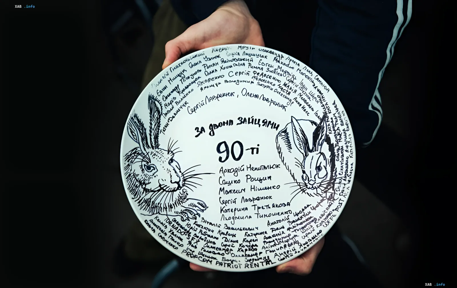 A person's hands hold a white plate with drawn hares and autographs of the cast and crew of Chasing Two Hares. The 90s
