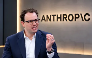 An Anthropic executive in a business suit gestures while speaking in front of the company logo