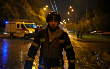 Rescuer in helmet and body armor against the background of nighttime Kyiv and emergency vehicles after shelling