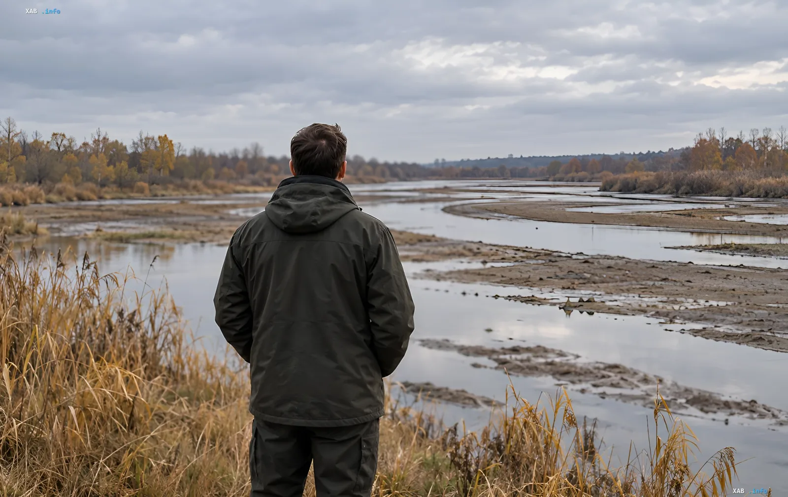 Man in a jacket looks at a shallow river with muddy banks under a cloudy sky