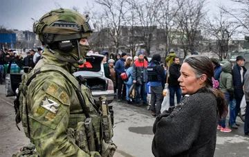 Озброєний російський військовослужбовець у шоломі з літерою Z перевіряє документи у жінки на вулиці під час рейдів в окупованому населеному пункті.