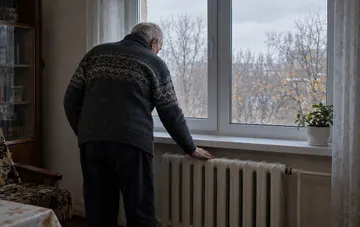 Літня людина біля вікна дивиться на вулицю біля опалювальної батареї в кімнаті