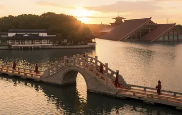 People in red attire walking across an arched bridge in a Chinese garden amid traditional architecture at sunset