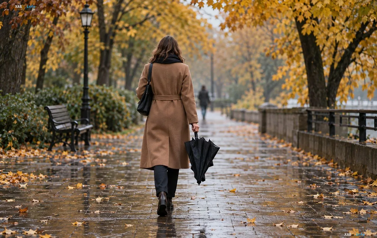 A woman in a coat with an umbrella walking down a park alley during an autumn rainy day