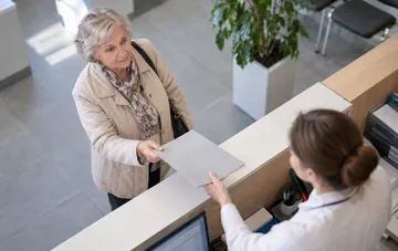 An elderly woman at the reception desk hands documents to a staff member