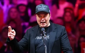 Elon Musk speaking on stage wearing a baseball cap during a public event