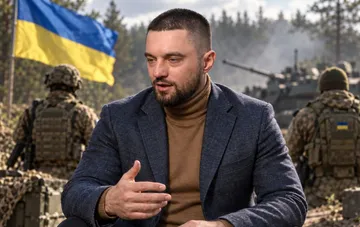 Украинский хореограф и победитель Танцев со звездами Макс Леонов в пиджаке и водолазке дает интервью на фоне украинского флага и военных
