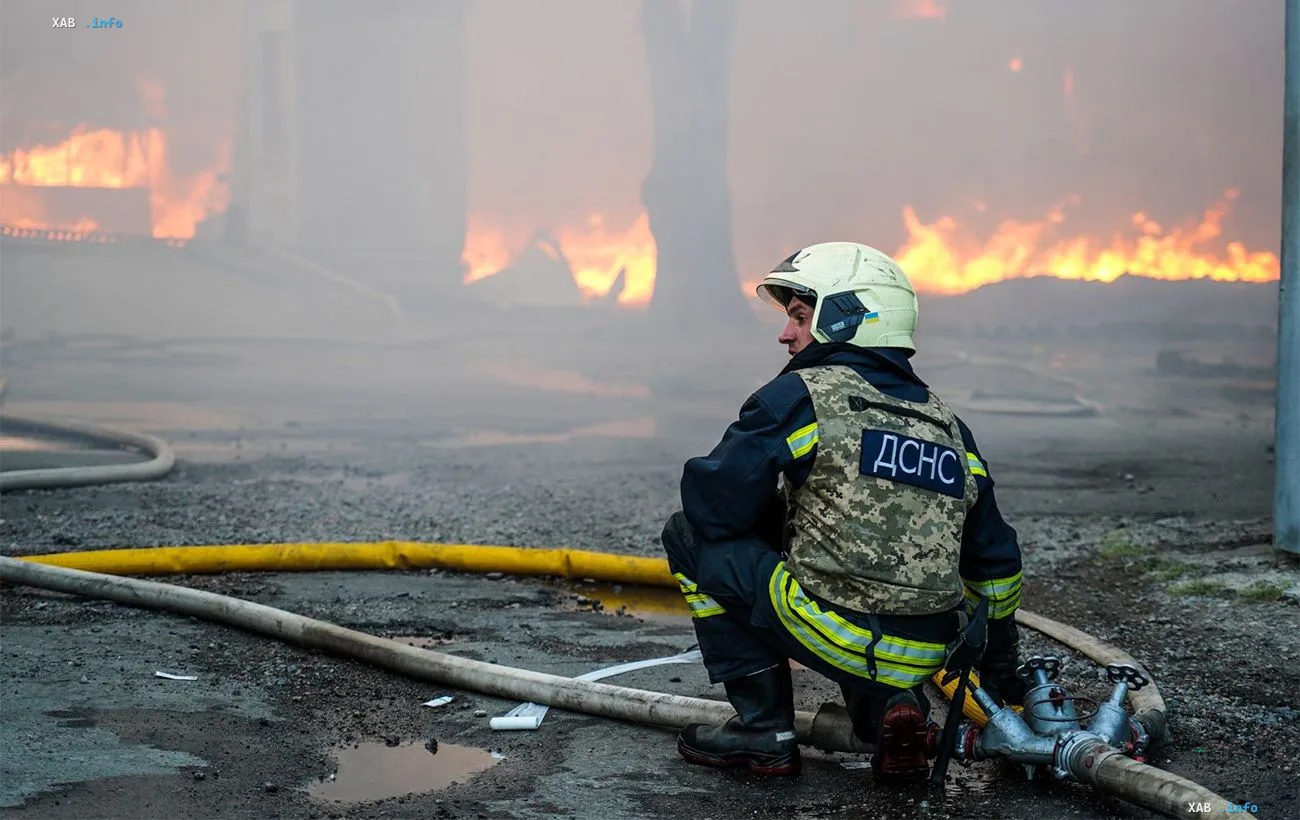 Рятувальник ДСНС у касці та захисному жилеті сидить на тлі масштабної пожежі після ракетного удару у Лозовій