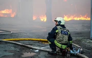 Спасатель ГСЧС в каме и защитном жилете сидит на фоне масштабного пожара и разрушений после ракетного удара в Лозовой