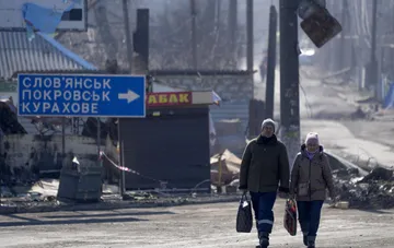 Люди идут по разрушенной улице на фоне указателя на Словянск, Покровск и Курахово