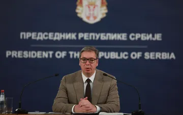 Бывший президент Сербии Александр Вучич выступает на пресс-конференции перед синим фоном с государственным гербом.