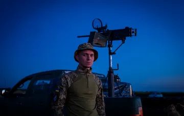 Украинский военнослужащий в камуфляжной панаме на фоне зенитной установки и пикапа в сумерках
