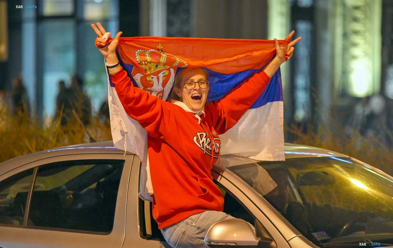 Женщина радостно машет сербским флагом, стоя в люке движущегося автомобиля в вечернее время