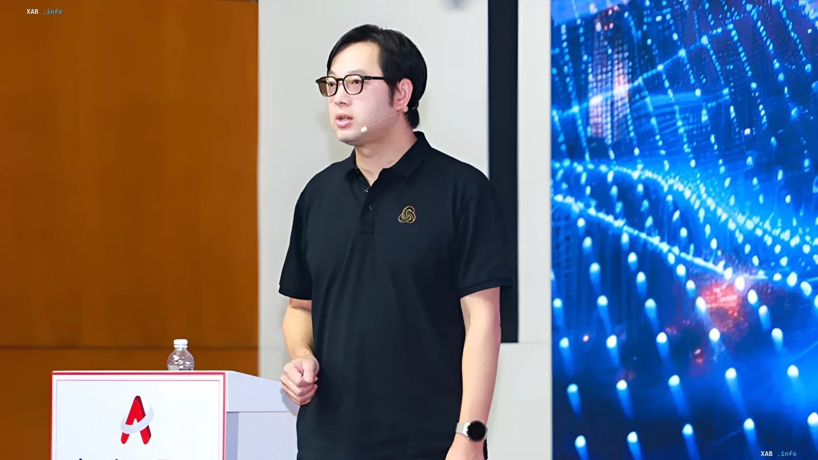Ein Mann in Brille und schwarzem Poloshirt spricht vor einem digitalen Bildschirm auf einer Technologiekonferenz