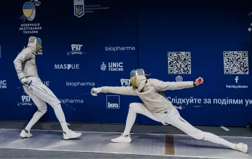 Две фехтовальщицы в защитных масках и белой форме соревнуются на официальной дорожке во время Кубка Украины