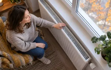 Eine Frau sitzt im Sessel und prüft mit der Hand einen warmen Heizkörper am Fenster