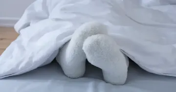 Weisse flauschige Socken an den Füssen unter einer weissen Decke im Bett