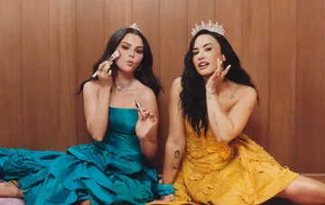 Selena Gomez und Demi Lovato mit Diademen und farbenfrohen Kleidern vor einer Holzwand während der Rare-Beauty-Kampagne
