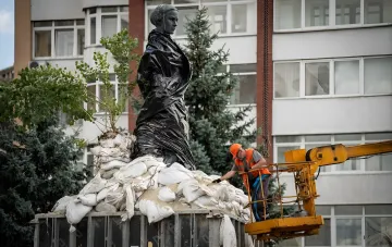 Рабочий в оранжевом жилете на подъемнике ремонтирует или проверяет укрытый черным полиэтиленом и мешками с песком памятник в городе