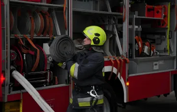Пожарный в форме и шлеме держит свернутый пожарный рукав на фоне специализированной пожарной машины.