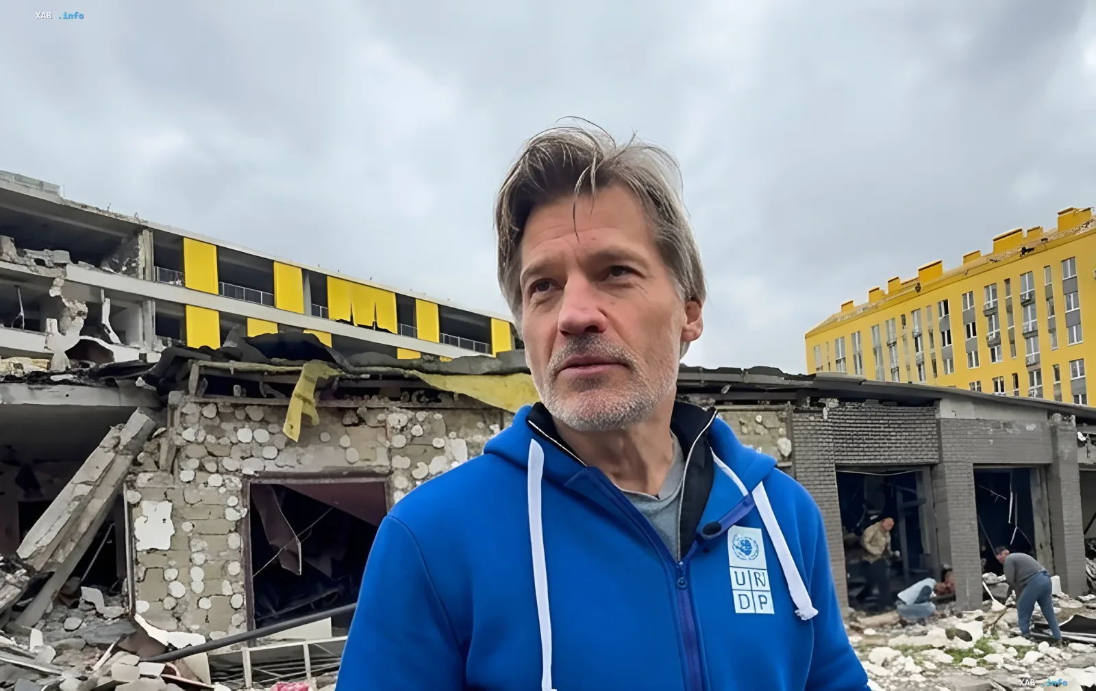 Danish actor Nikolaj Coster-Waldau in a blue UNDP hoodie against the backdrop of a shell-damaged residential building in Kyiv