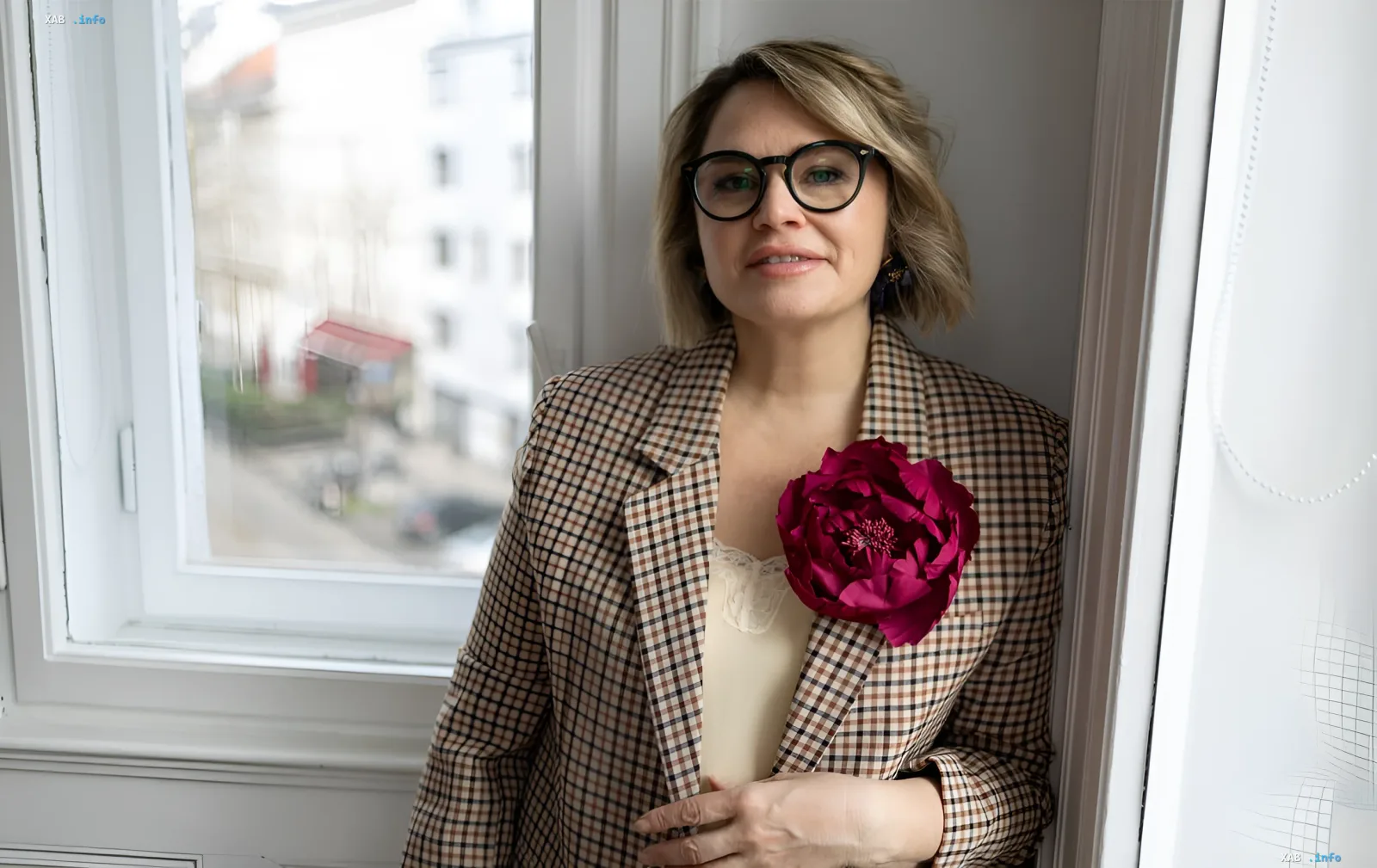Woman in glasses and a plaid blazer with a flower brooch by the window