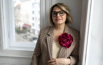 Frau mit Brille und kariertem Blazer mit Blumenbrosche am Fenster