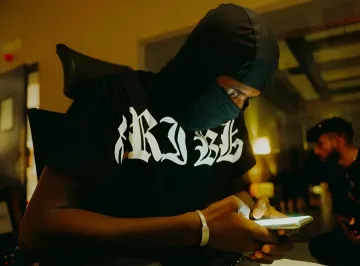 A person in a balaclava sits indoors checking their smartphone under yellow lighting