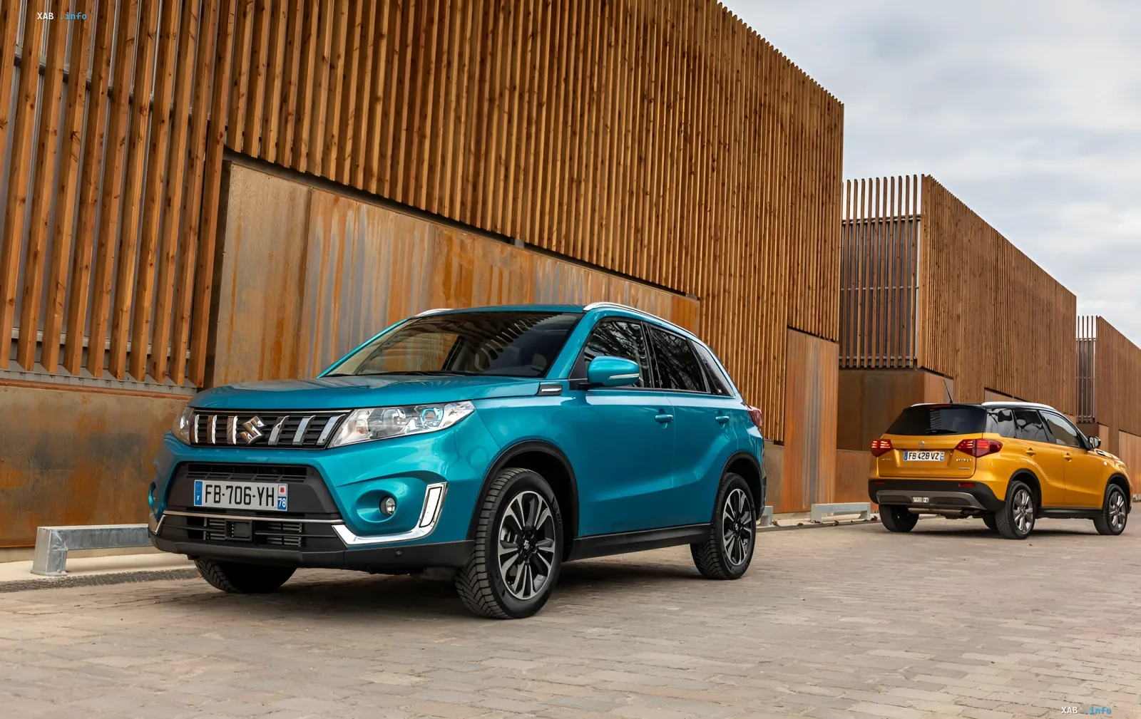 Бирюзовый и желтый кроссоверы Suzuki Vitara припаркованы на фоне современного здания с деревянной отделкой